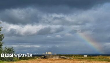 Plenty of cloud and a rainbow over East Lothian, Scotland. There is yellow grass in the foreground and tall cream-coloured buildings in the distance.