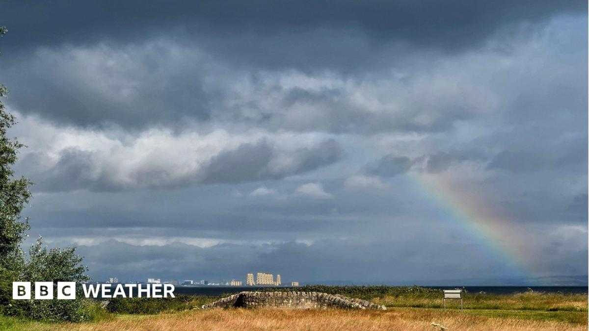 Plenty of cloud and a rainbow over East Lothian, Scotland. There is yellow grass in the foreground and tall cream-coloured buildings in the distance.