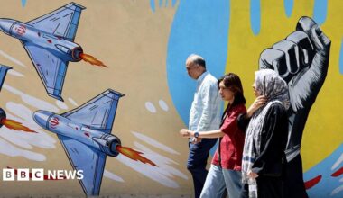 An older man and woman and a younger woman walking past a mural in Tehran featuring several drones and a raised fist
