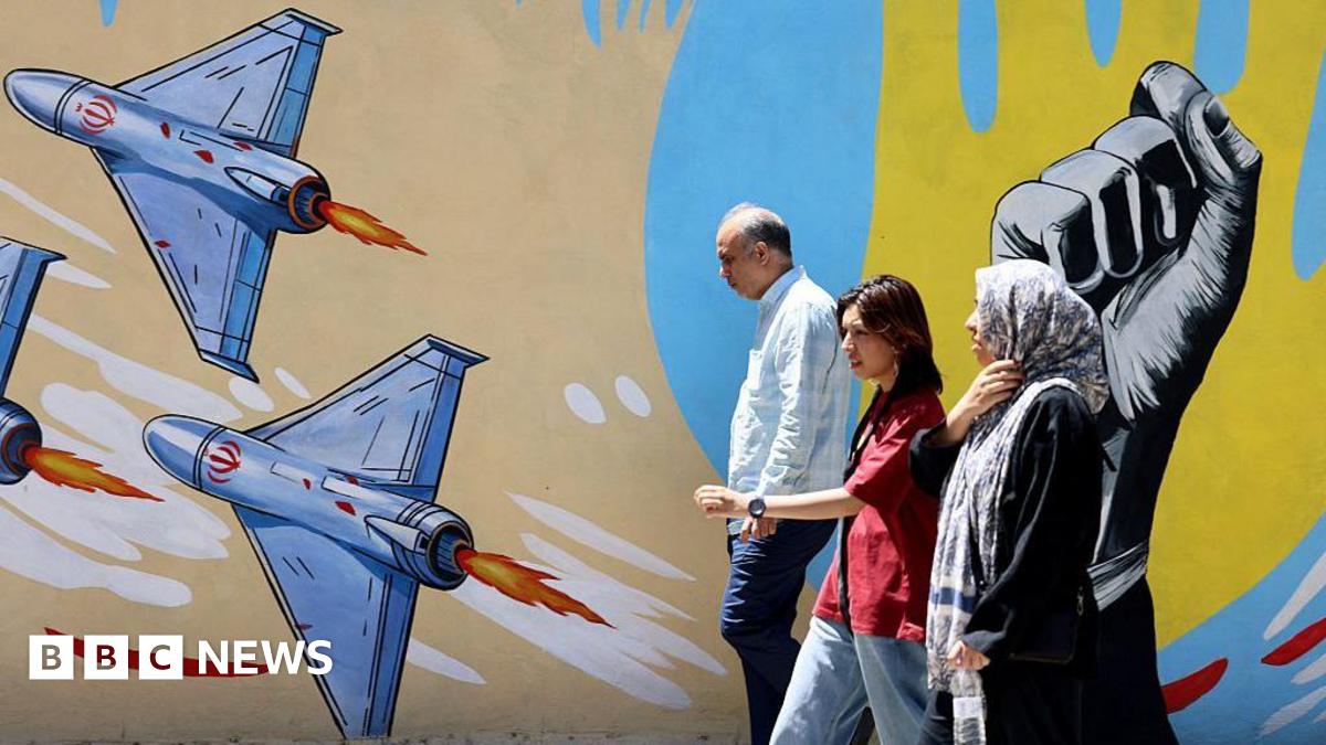 An older man and woman and a younger woman walking past a mural in Tehran featuring several drones and a raised fist