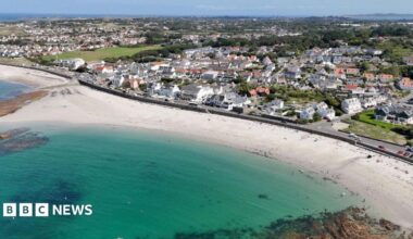 A drone picture showing Cobo bay in Guernsey. Blue water against white sandy beach with buildings and roads in the background.