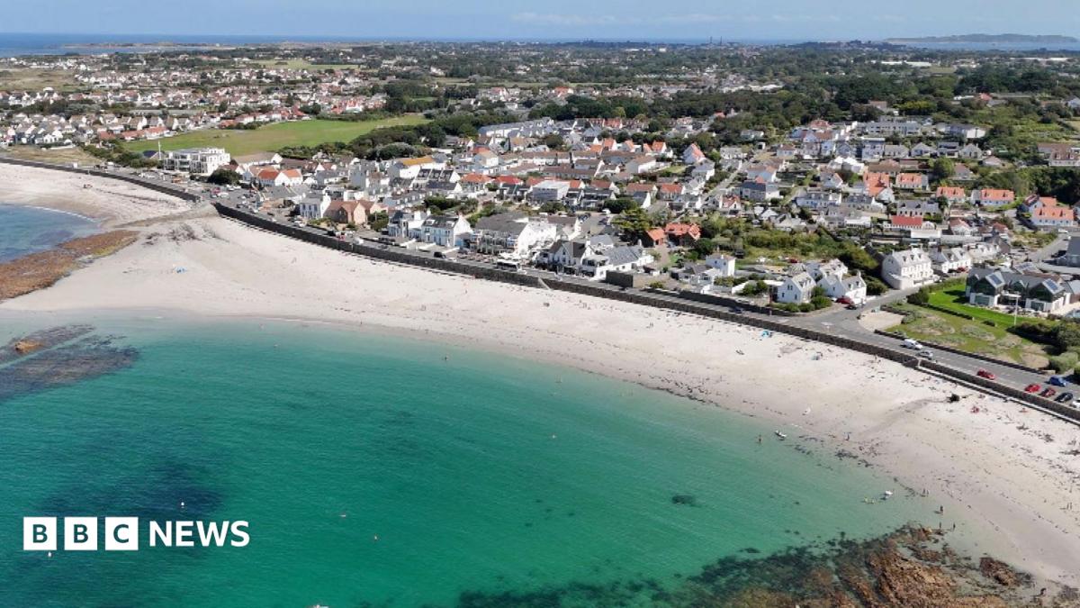 A drone picture showing Cobo bay in Guernsey. Blue water against white sandy beach with buildings and roads in the background.
