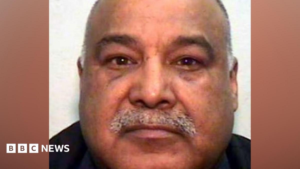 A mugshot of an Asian man in his 60s, with a grey moustache and bald head. He is looking into the camera.