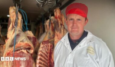 A man in a large white coat and a red hat stood in front of butchered meat hanging from a ceiling
