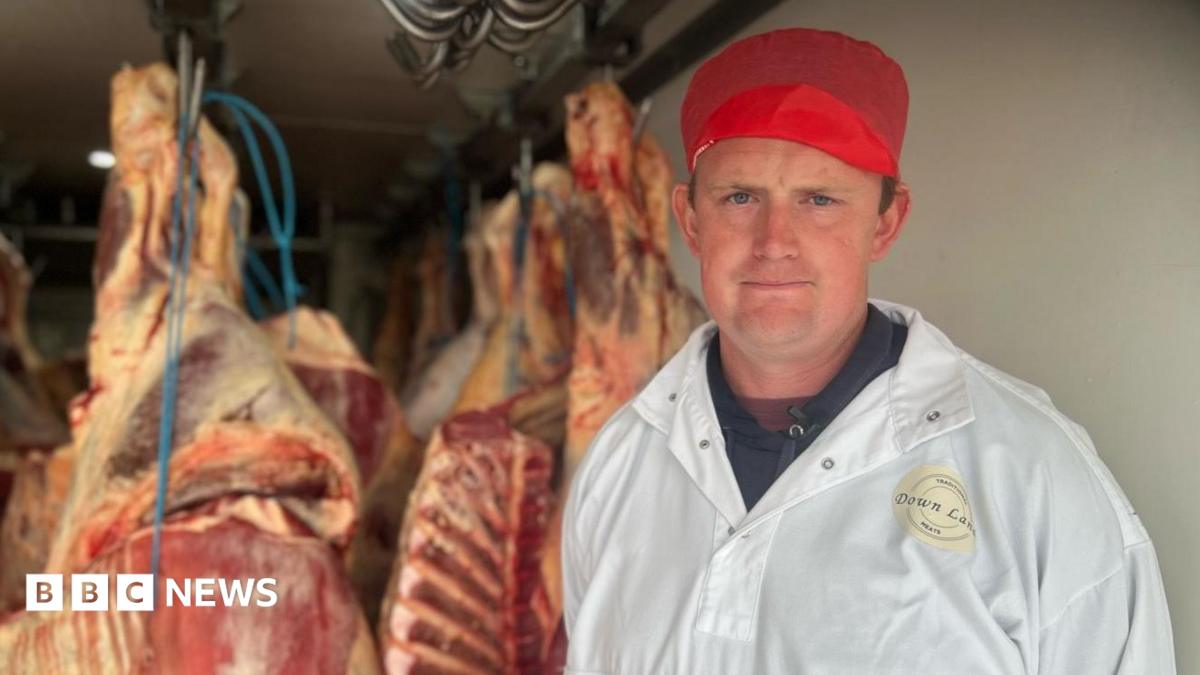 A man in a large white coat and a red hat stood in front of butchered meat hanging from a ceiling