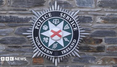 The official badge of the PSNI. It is hanging on a grey brick wall. It has a red x in the middle and a number of other crime and justice related symbols on the outside. The include: a crown, weighing scales and a torch.