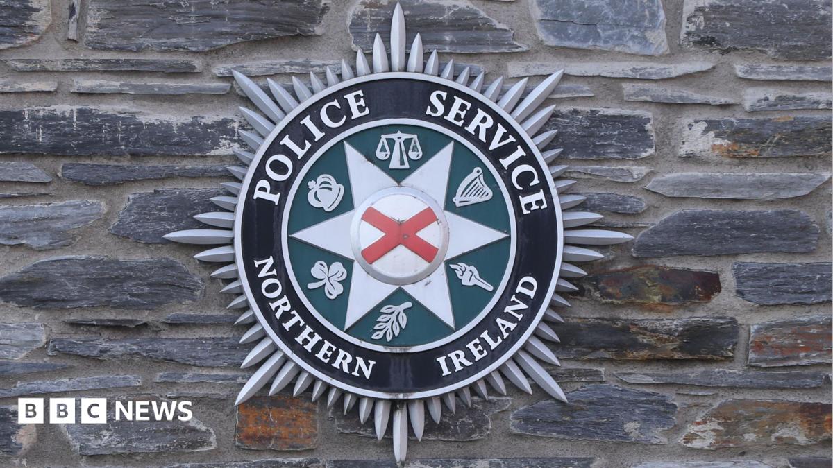The official badge of the PSNI. It is hanging on a grey brick wall. It has a red x in the middle and a number of other crime and justice related symbols on the outside. The include: a crown, weighing scales and a torch.