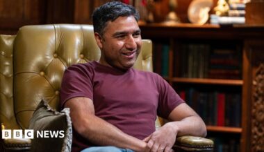 Nick Mohammed is sitting in a large leather armchair. He is a youngish man with dark hair and a bit of stubble and wearing a dark red top with jeans.