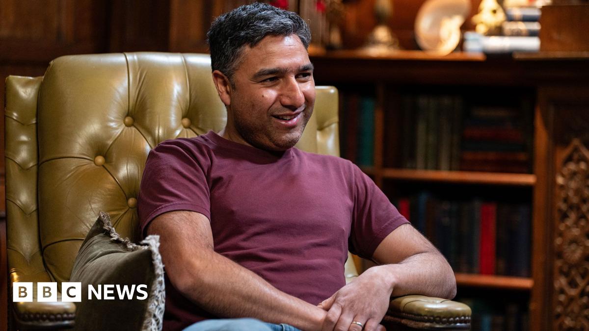 Nick Mohammed is sitting in a large leather armchair. He is a youngish man with dark hair and a bit of stubble and wearing a dark red top with jeans.