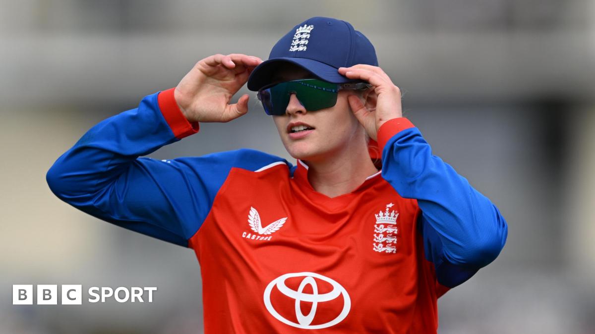 England Women's stand-in captain Charlie Dean stands in the outfield during a game, wearing a blue England cap and black sunglasses
