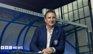 Robert Smethurst wears a navy blue suit with a club logo on the pocket. He wears a white shirt and stands pitch side in front of a a Macclesfield Football Club Home stand.