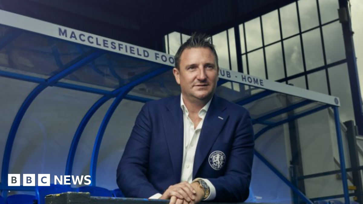 Robert Smethurst wears a navy blue suit with a club logo on the pocket. He wears a white shirt and stands pitch side in front of a a Macclesfield Football Club Home stand.