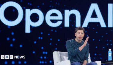 Sam Altman sits in a chair on a stage. He is motioning with his hands. Behind him is the Open AI logo.