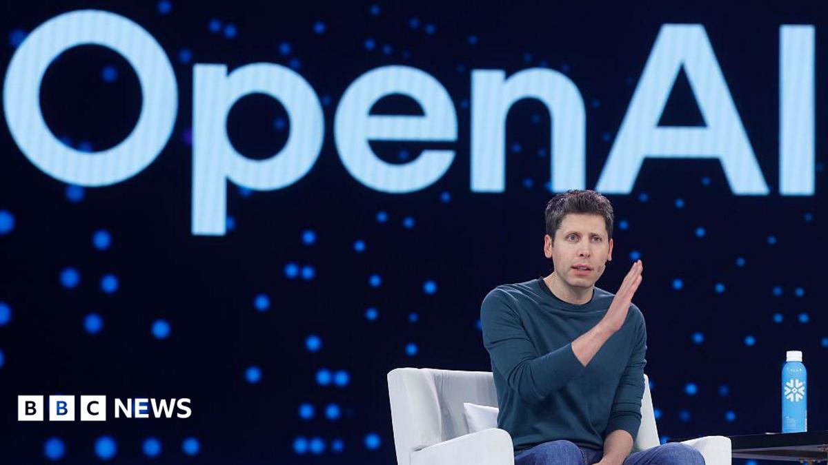 Sam Altman sits in a chair on a stage. He is motioning with his hands. Behind him is the Open AI logo.