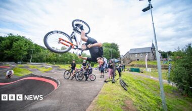 A bike rider is upside down over a track with other riders looking on and a large granite building in the background.