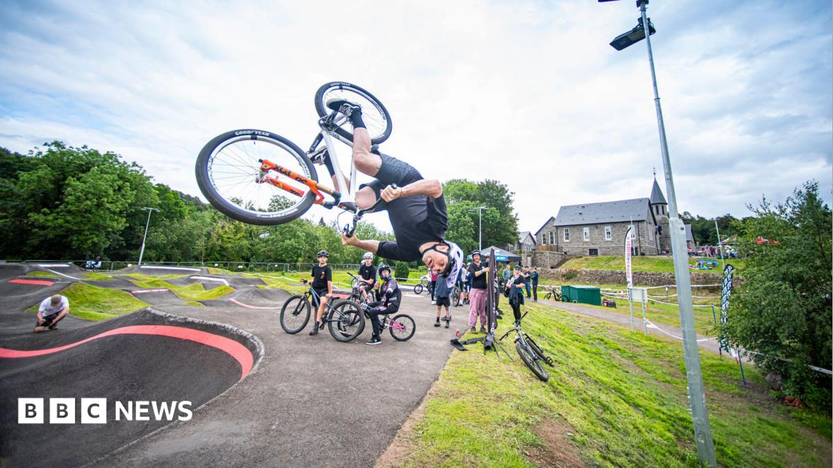 A bike rider is upside down over a track with other riders looking on and a large granite building in the background.