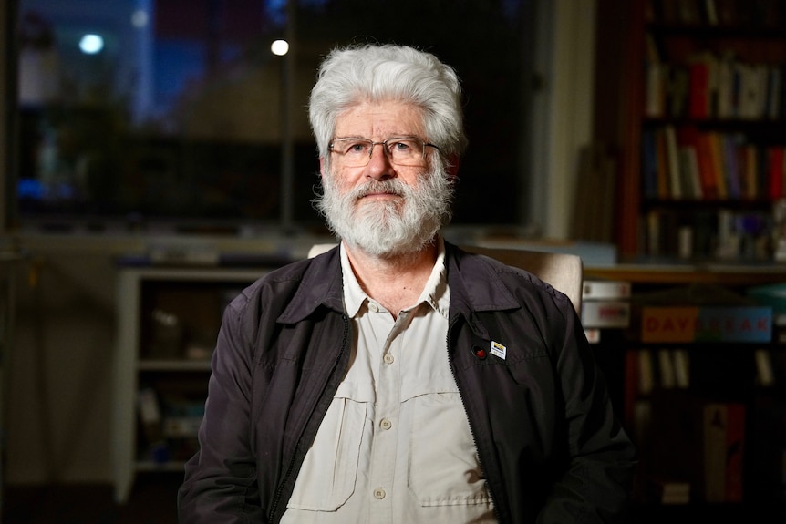 Man in a grey shirt and black jacket. He has a beard and wears glasses.
