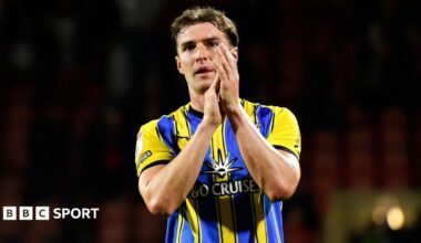 James Bree claps fans with hands to side of his face, wearing blue and yellow away kit