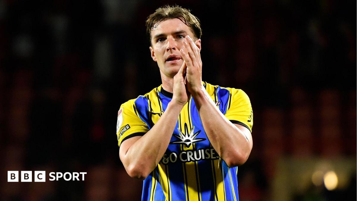 James Bree claps fans with hands to side of his face, wearing blue and yellow away kit