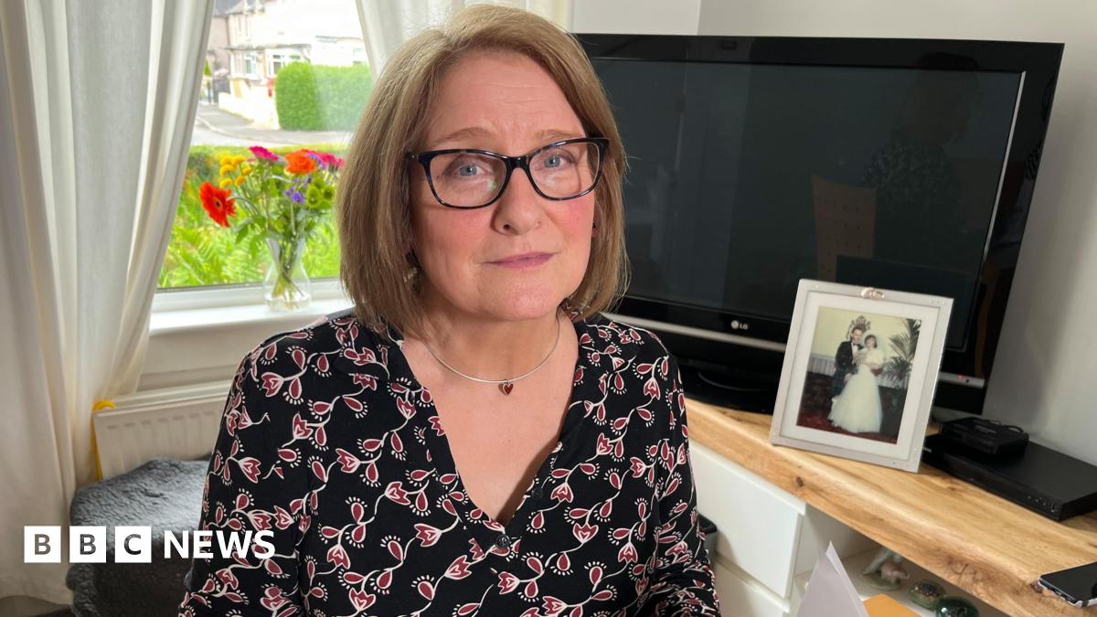 Kay Donald is a woman in her 60s with light brown shoulder-length hair and dark-rimmed glasses. She is wearing a black and red patterned top and is sitting in front of a large TV and window which looks out onto a garden with a street beyond.