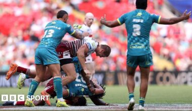 Wigan Warriors player Sam Walters, in cherry and white hoops, dumps Hull Kingston Rovers player Bill Leyland on the ground head-first, in a tackle made with team-mate Junior Nsemba, partly hidden. Hull KR players Eliot Minchella and Peta Hiku look on.