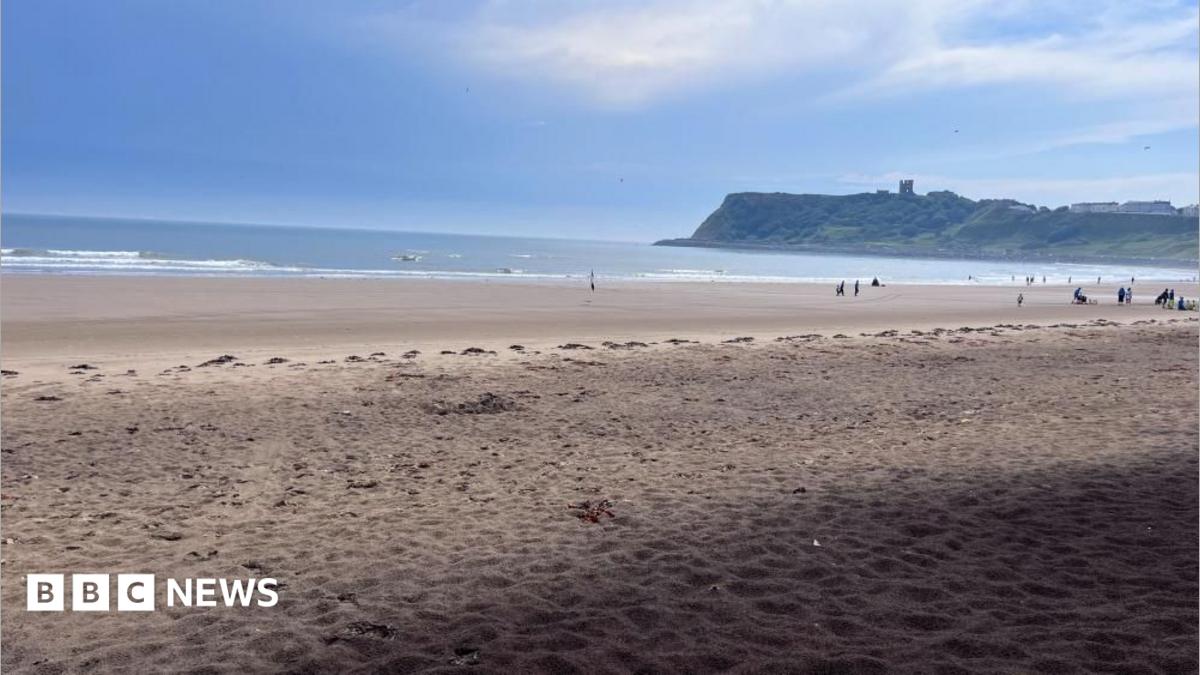 A view of North Bay in Scarborough, a stretch of sandy beach with the sea in the background. In the distance, a ruined castle can be seen on cliff tops.