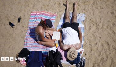 Two people sat on a sandy beach. A woman is sat up reading while a man is led out on a towel next to her. Both of them are wearing hats.