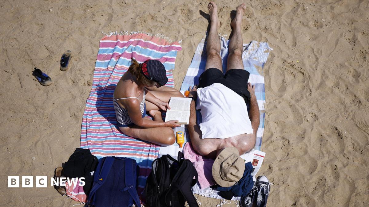 Two people sat on a sandy beach. A woman is sat up reading while a man is led out on a towel next to her. Both of them are wearing hats.
