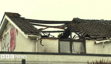The roof of a building after a fire. Large parts of the roof is missing. The sky is grey and cloudy. Old graffiti is on the side of the building.