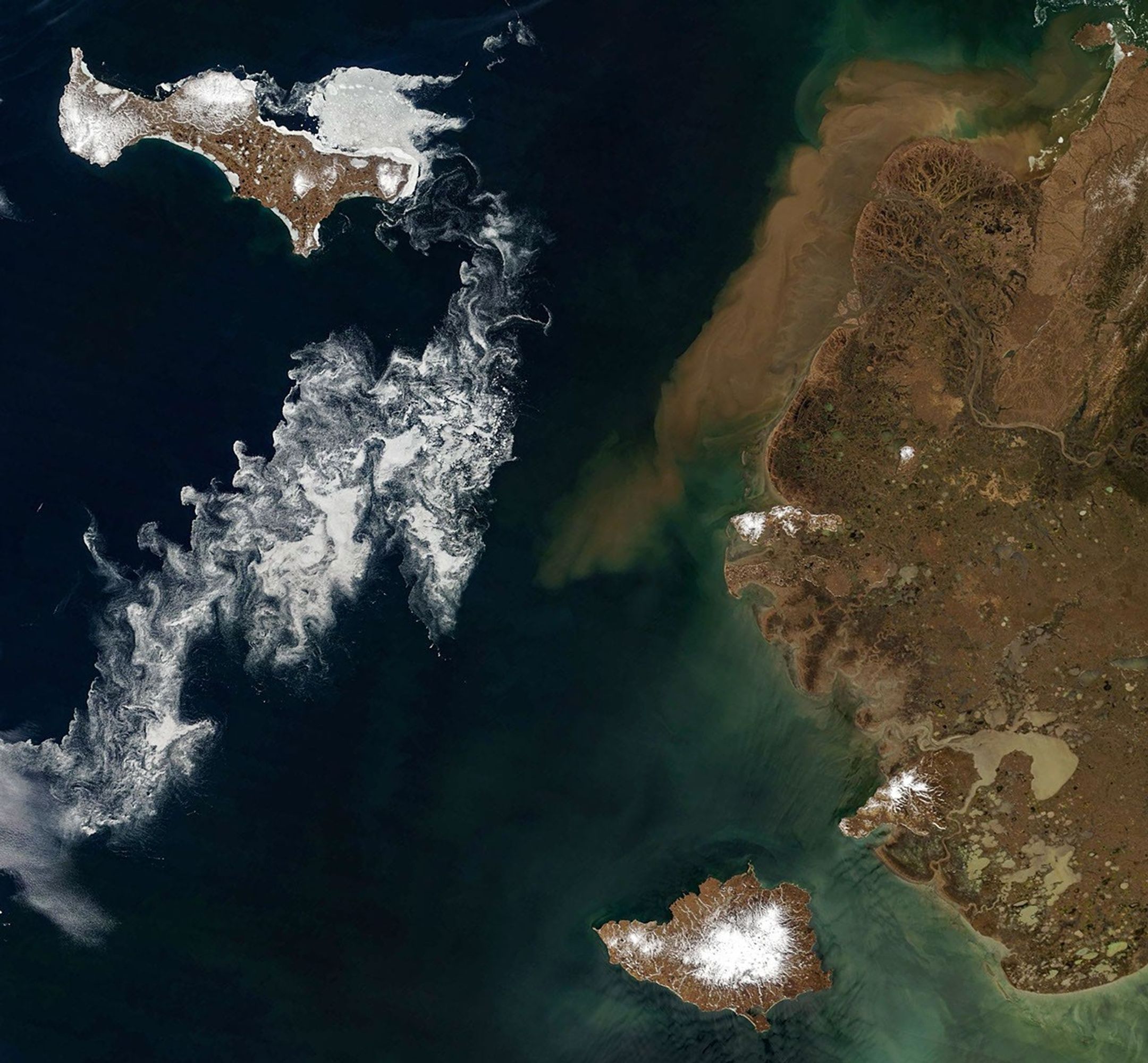 Two large, partly snow-covered islands lie west of mainland Alaska. Sea ice fragments form swirling patterns in the ocean, and brown water lines part of the Alaskan coastline.