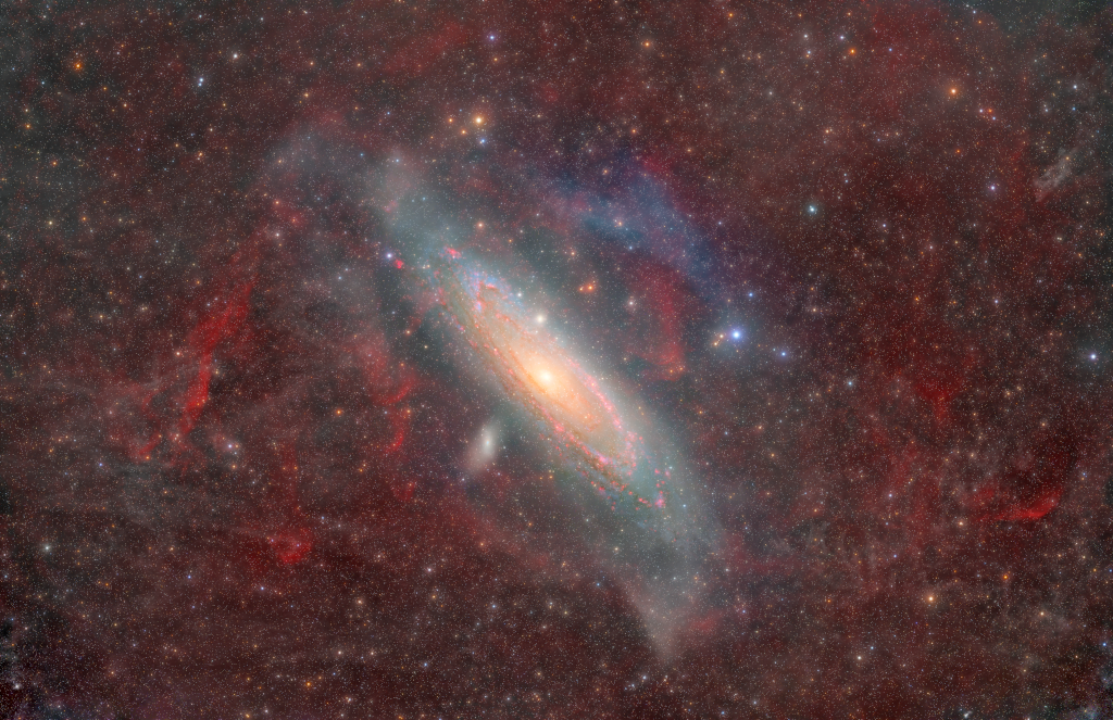 Wispy clouds of dust and gas in the Milky Way obscure the image. Milky Way stars are scattered across the image. Andromeda is a tight spiral of gas, dust, and stars that 
occupies the middle background. A couple smaller galaxies look like small bright clumps of stars near Andromeda.