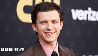 Male actor Tom Holland, wearing a pink shirt, black tie and brown jacket, smiling.