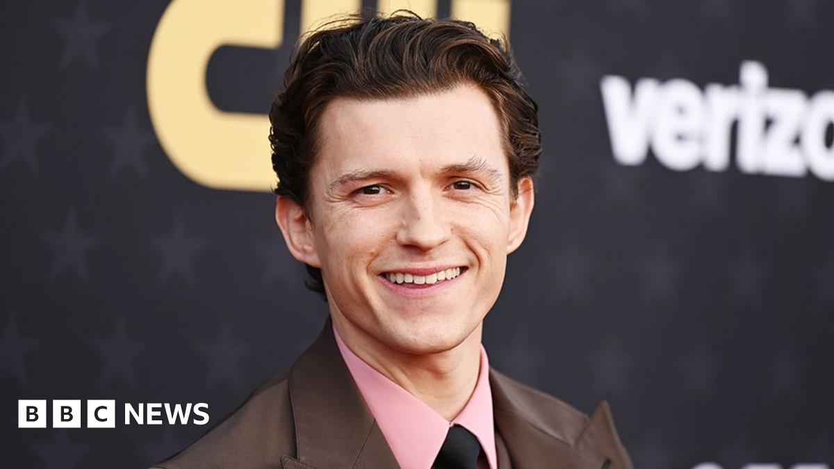 Male actor Tom Holland, wearing a pink shirt, black tie and brown jacket, smiling.