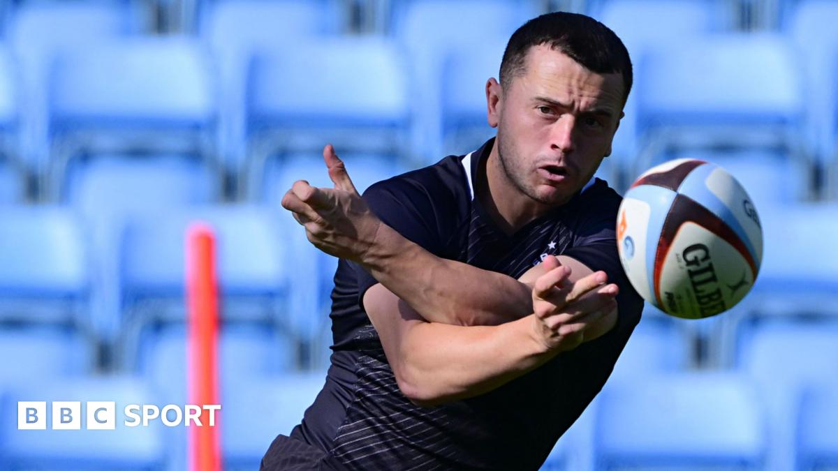 Charlie Chapman spins a pass out of his left hand sending the ball straight at the viewer. He watches it closely as it flies away from him. In the background there are empty light blue seats which are blurred and the top part of a red training stick.
