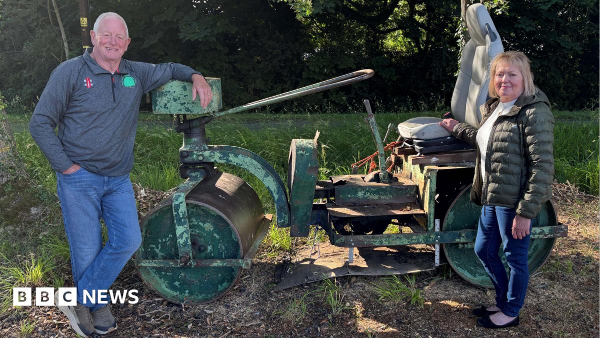 David wears a grey top with the Battisford & District club logo on it, while Julia wears a green jacket and denim jeans. They are standing at either end of a green old cricket pitch roller, in front of some trees.