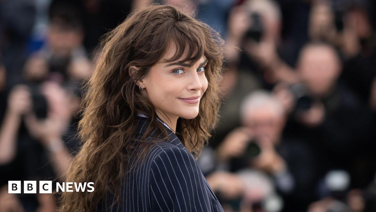 Cara Delevingne wears a navy pinstripe jacket. She is looking over her shoulder and has brown wavy hair.