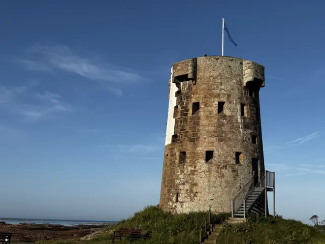 A picture of Le Hocq Tower. It is a brick building with stairs to the left of it. There is a body of water in the distance.
