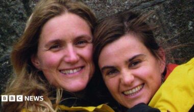 Jo Cox MP, a brunette woman, smiles at the camera. She wears an orange top with a necklace. Behind her the Houses of Parliament are blurred.