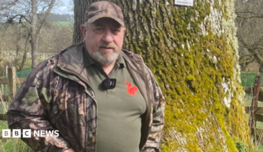 Ian Glendinning is a man in his 60s. He is standing in front of a tree wearing a camouflage jacket and a green top with a red squirrel logo on it.