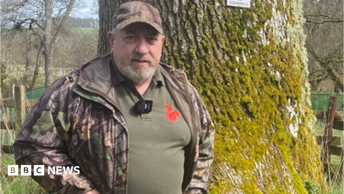 Ian Glendinning is a man in his 60s. He is standing in front of a tree wearing a camouflage jacket and a green top with a red squirrel logo on it.