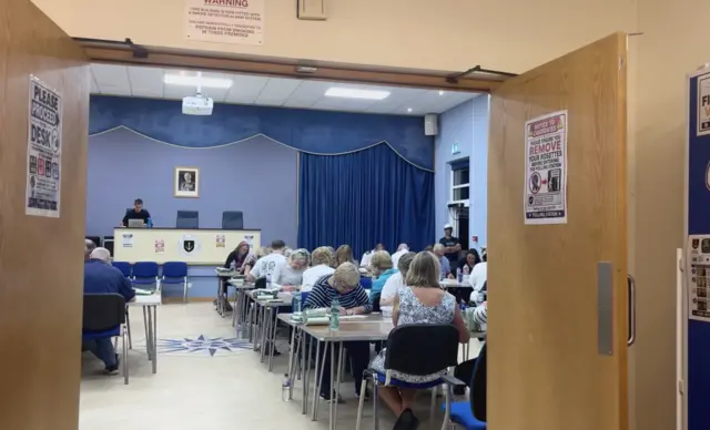 A picture of St Clement Parish Hall. There is a number of people inside the hall counting.