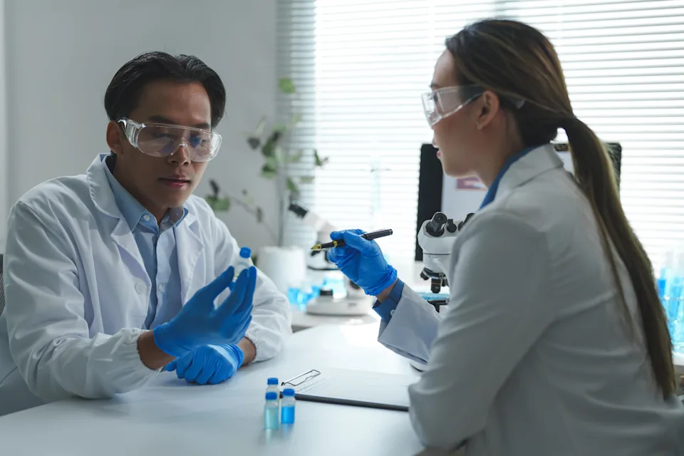 Two scientists in a lab discussing a small vial. Both are wearing lab coats, safety goggles, and gloves in a research setting