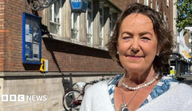 Joanna Brittan is looking at the camera and smiling. She has a short dark bob and is wearing a pearl necklace and earrings. She wears a white fluffy cardigan and is pictured from the shoulders up. She is standing outside a police station.