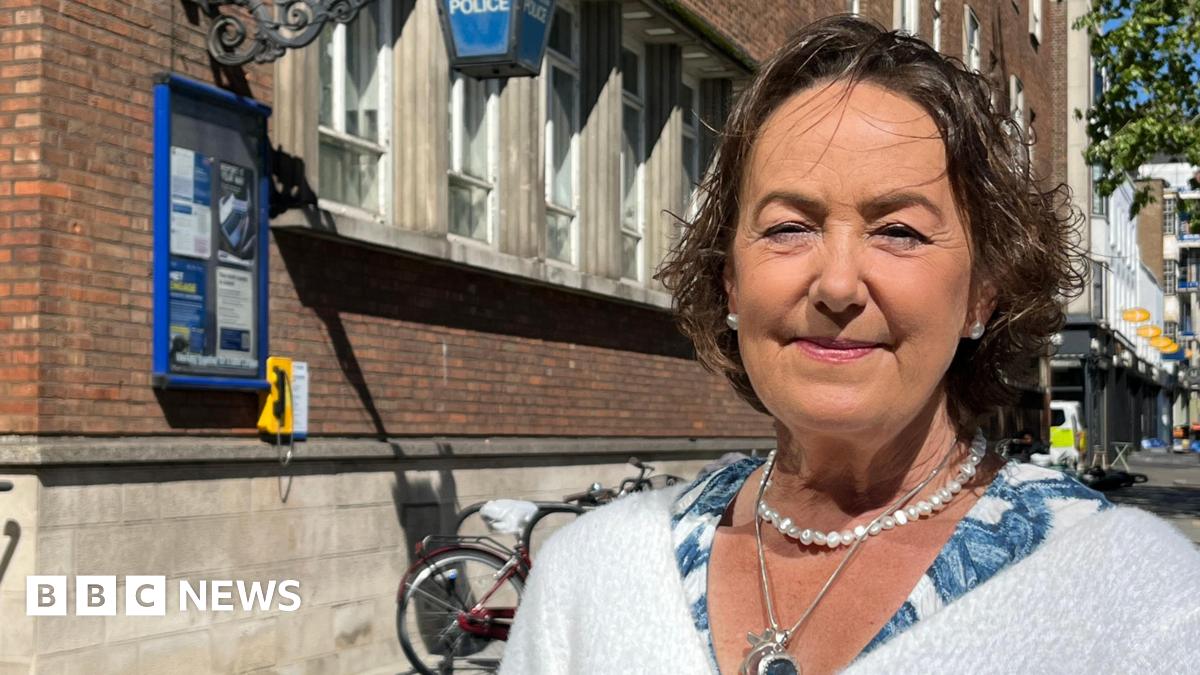 Joanna Brittan is looking at the camera and smiling. She has a short dark bob and is wearing a pearl necklace and earrings. She wears a white fluffy cardigan and is pictured from the shoulders up. She is standing outside a police station.