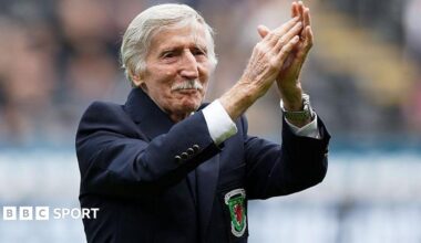 Cliff Jones, who has white short hair and a moustache, has his arms raised clapping. He has a blue blazer on with the Wales logo.