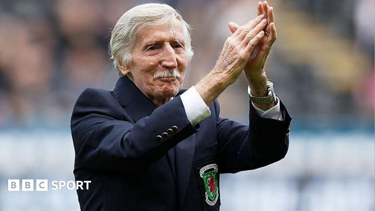 Cliff Jones, who has white short hair and a moustache, has his arms raised clapping. He has a blue blazer on with the Wales logo.