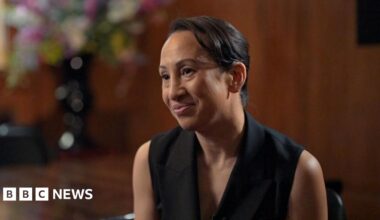 Gleit wears a sleeveless black blazer, and is sitting on a chair in front of a large glossy wood panelled room. A large vase of colourful flowers is also blurred in the background behind her.