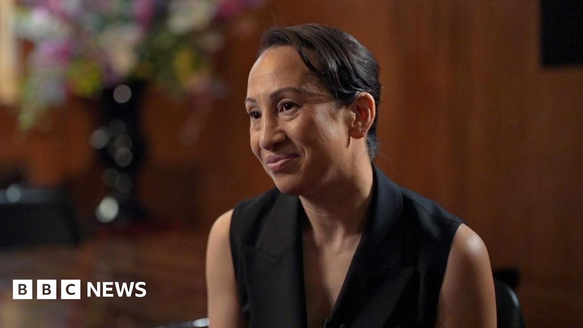 Gleit wears a sleeveless black blazer, and is sitting on a chair in front of a large glossy wood panelled room. A large vase of colourful flowers is also blurred in the background behind her.