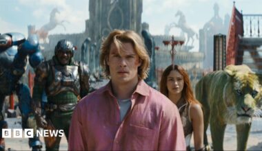 Masters of the Universe still. A group of superhero characters and a CGI lion are stood in a line in front of a tall building.