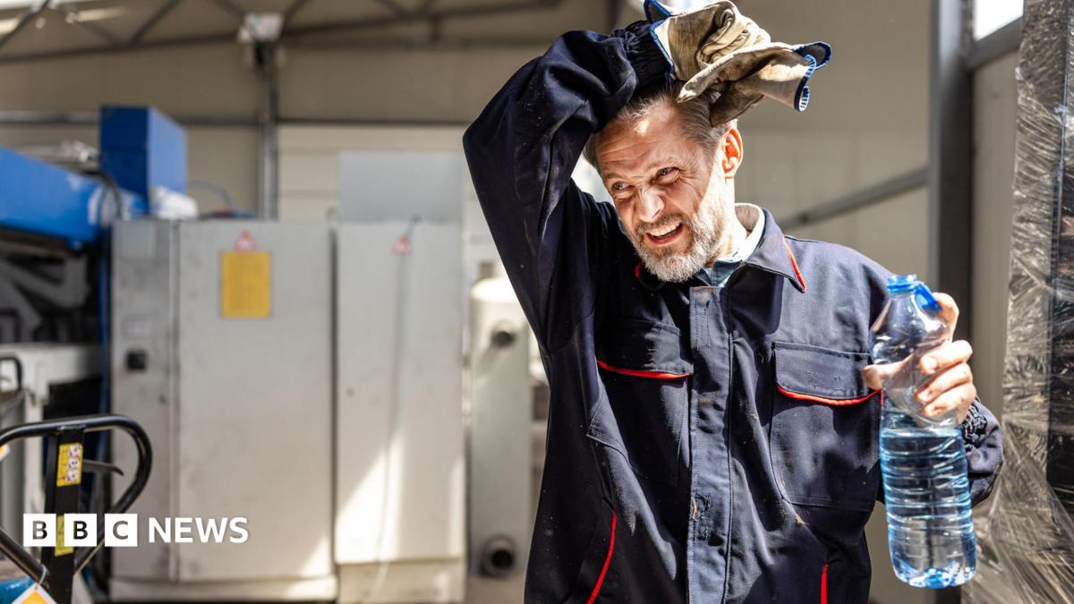 A man in overalls wiping his forehead and holding a plastic water bottle